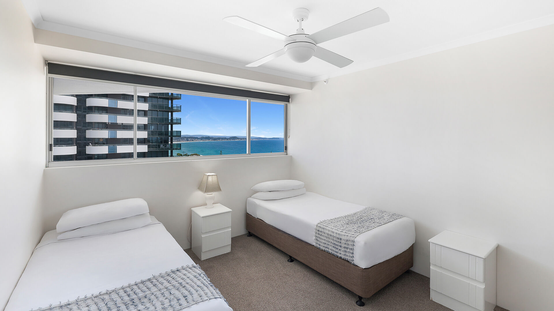 Bright twin bedroom with two single beds, white bedside tables, a ceiling fan, and a large window offering ocean and city views.