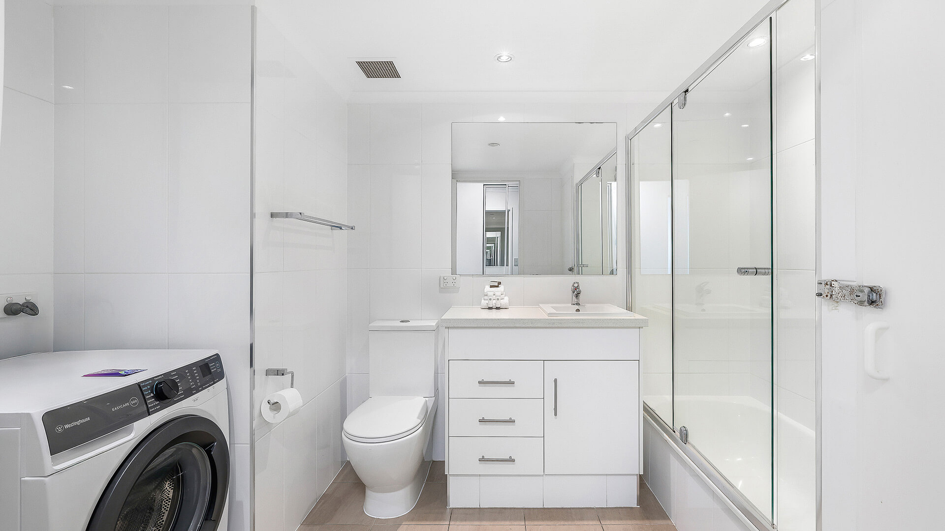 Bright modern bathroom with a bathtub-shower combo, white vanity, toilet, and an in-room front-load washing machine.