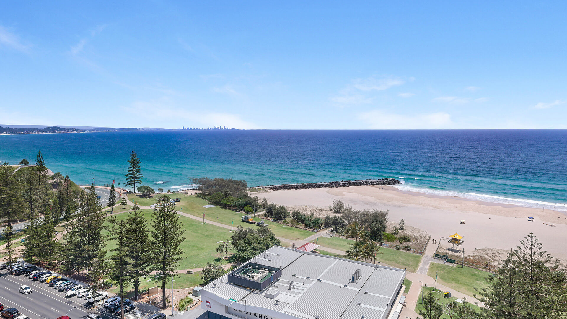 Aerial-style view of Coolangatta Beach & foreshore parklands, showing the Coolangatta Surf Life Saving Club, ocean & distant skyline on the horizon.