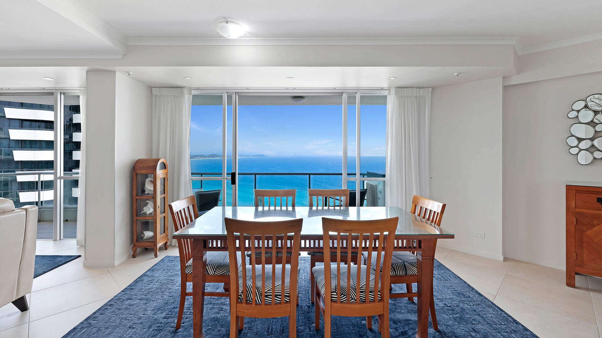 Dining area of a Superior 3 Bedroom Ocean View Apartment featuring wooden dining table & chairs, with sliding doors opening to panoramic ocean views.