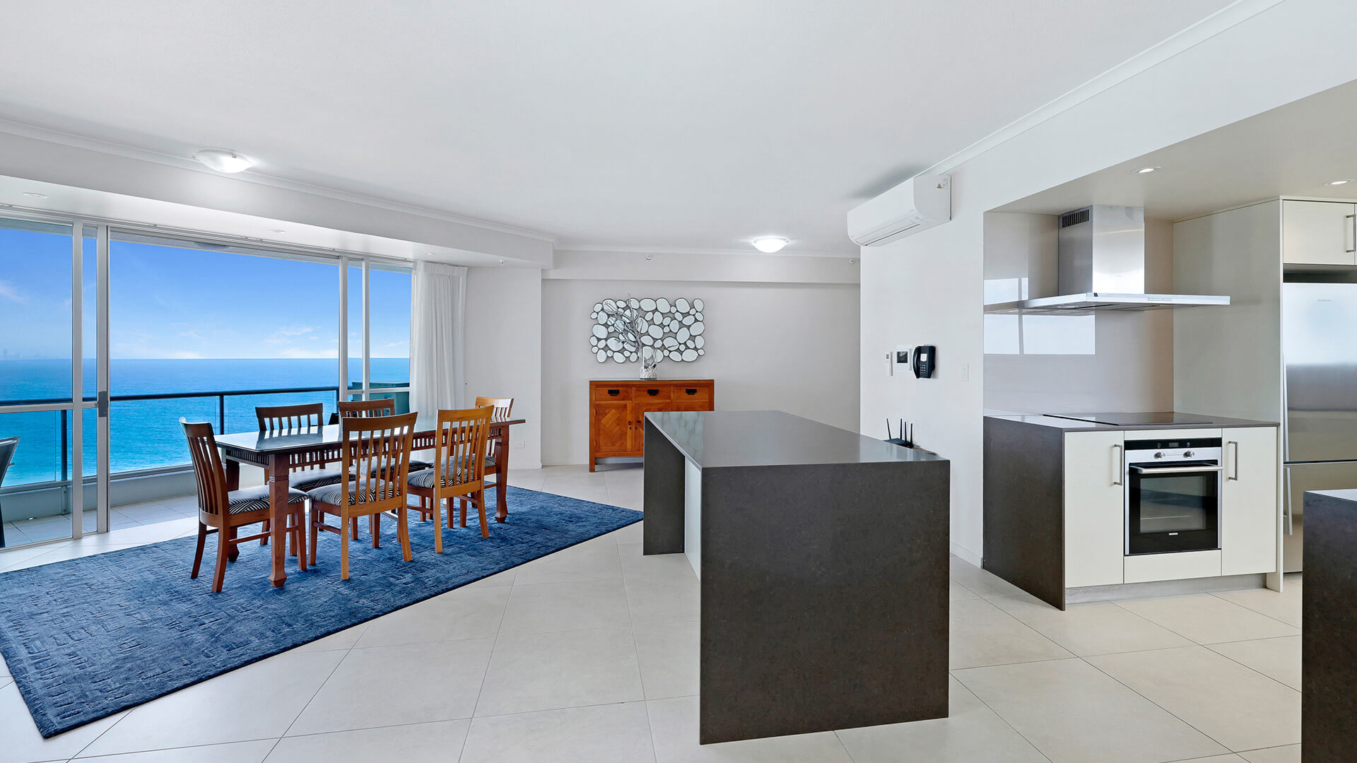 Open-plan dining & kitchen area in a Superior 3 Bedroom Ocean View Apartment featuring large island bench & wooden dining setting with ocean views.