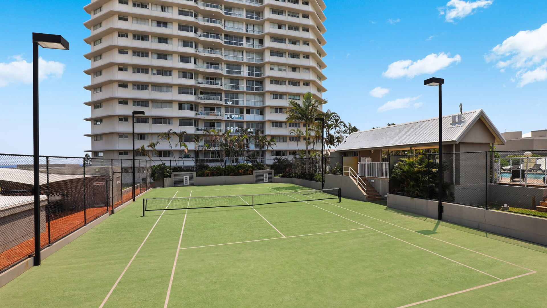 Outdoor tennis court at ULTIQA Points North with a high-rise backdrop, surrounded by fencing and landscaped gardens under a bright blue sky.