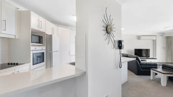 Open-plan layout with modern white kitchen, built-in appliances and breakfast bar, looking into the living area with leather seating and a TV.