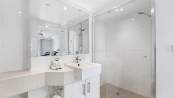 Bright modern bathroom with a large mirror, white vanity, glass-enclosed walk-in shower, and neatly folded towels on the counter.
