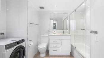 Bright modern bathroom with a bathtub-shower combo, white vanity, toilet, and an in-room front-load washing machine.