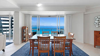 Dining area of a Superior 3 Bedroom Ocean View Apartment featuring wooden dining table & chairs, with sliding doors opening to panoramic ocean views.