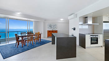 Open-plan dining & kitchen area in a Superior 3 Bedroom Ocean View Apartment featuring large island bench & wooden dining setting with ocean views.