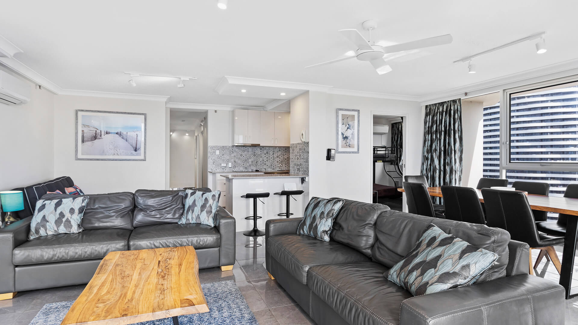 Living room area with a black leather couch, blue rug, wooden coffee table and kitchen in the background at ULTIQA Beach Haven.
