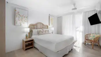 Coastal-style featuring a queen bed with rattan headboard, neutral tones, natural light, and a wall-mounted TV.