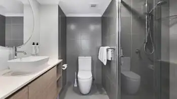 Modern bathroom featuring a walk-in glass shower, toilet, floating vanity with vessel sink, round mirror, and tiled walls in soft grey and white tones.