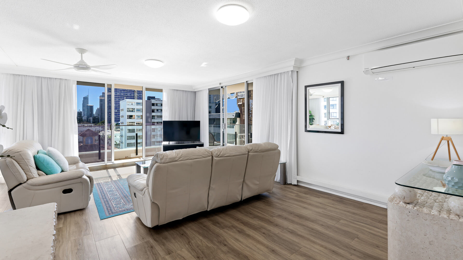 Spacious living room with leather lounges and flat-screen TV in ULTIQA Beach Haven’s Standard 2 Bedroom Ocean View Apartment, featuring large windows and city views.