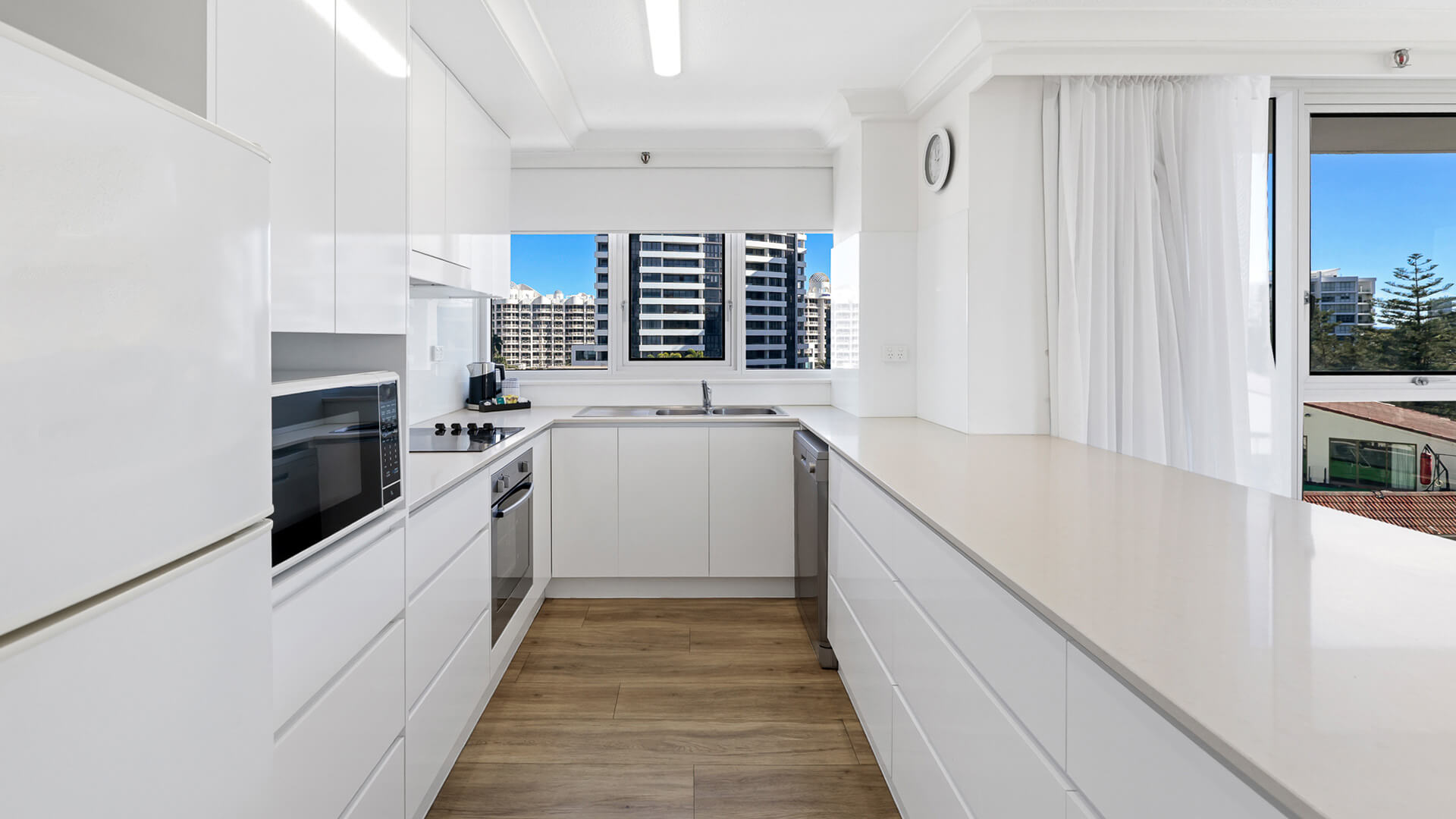 Modern kitchen in the Standard 2 Bedroom Ocean View Apartment at ULTIQA Beach Haven, featuring sleek white cabinetry, full-size appliances, and city views through large windows.