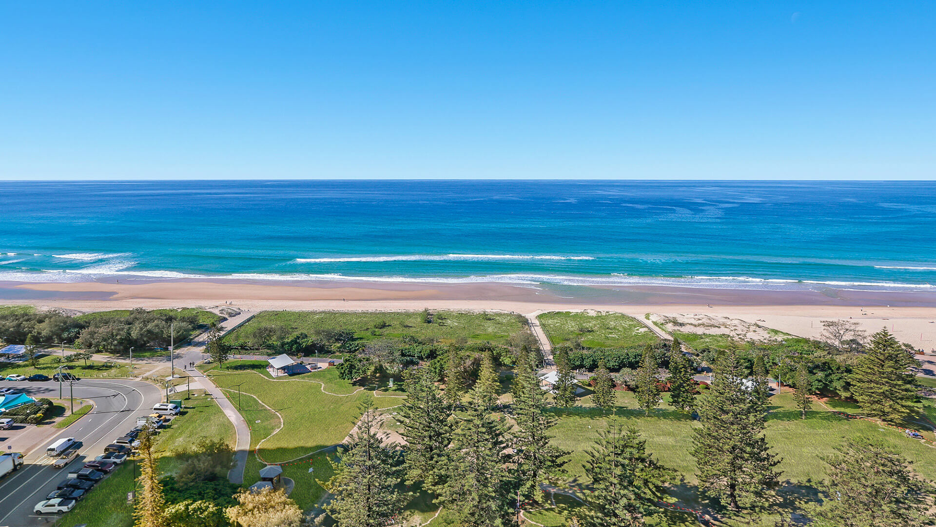 Aerial view of parklands and pathways leading to the beach, with clear blue skies and waves rolling onto the sand, seen from the 2 Bedroom Ocean View Apartment at ULTIQA Beach Haven.