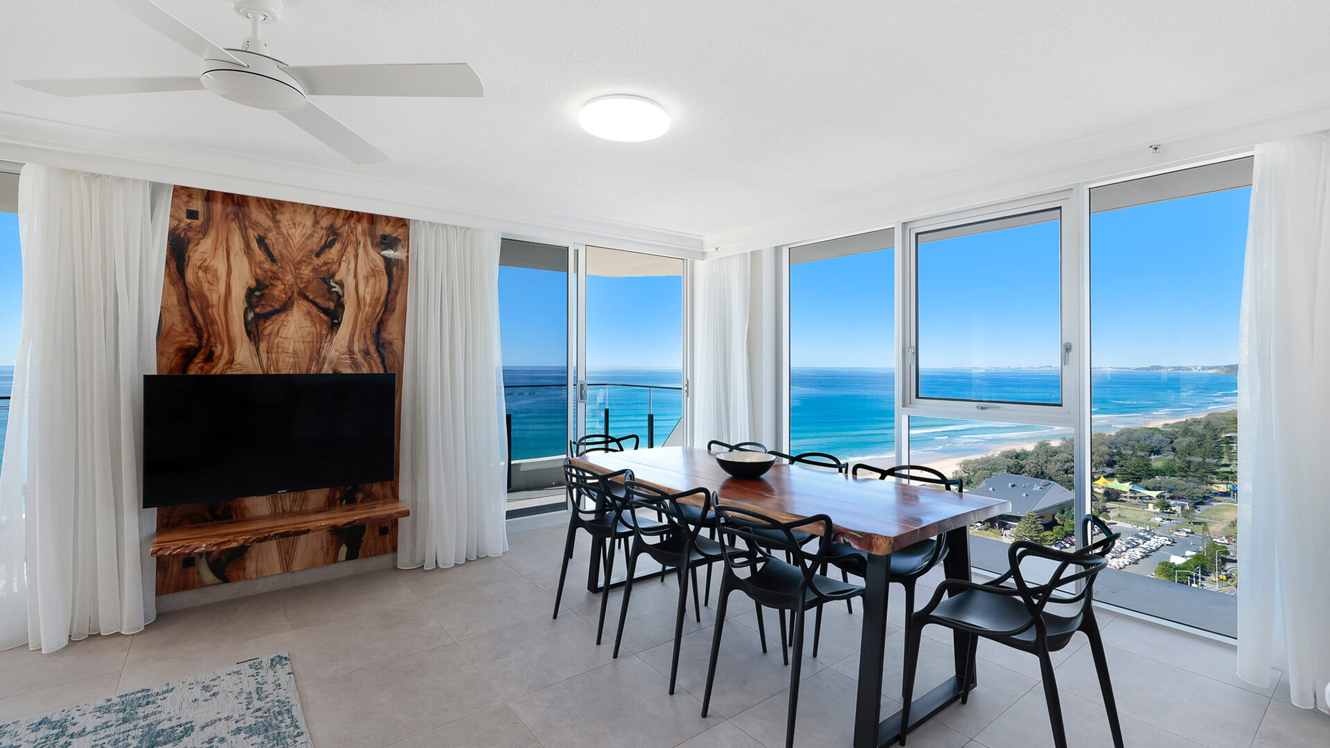 Dining area featuring a wooden dining table, modern black chairs, and panoramic views of the Gold Coast coastline through floor-to-ceiling windows.