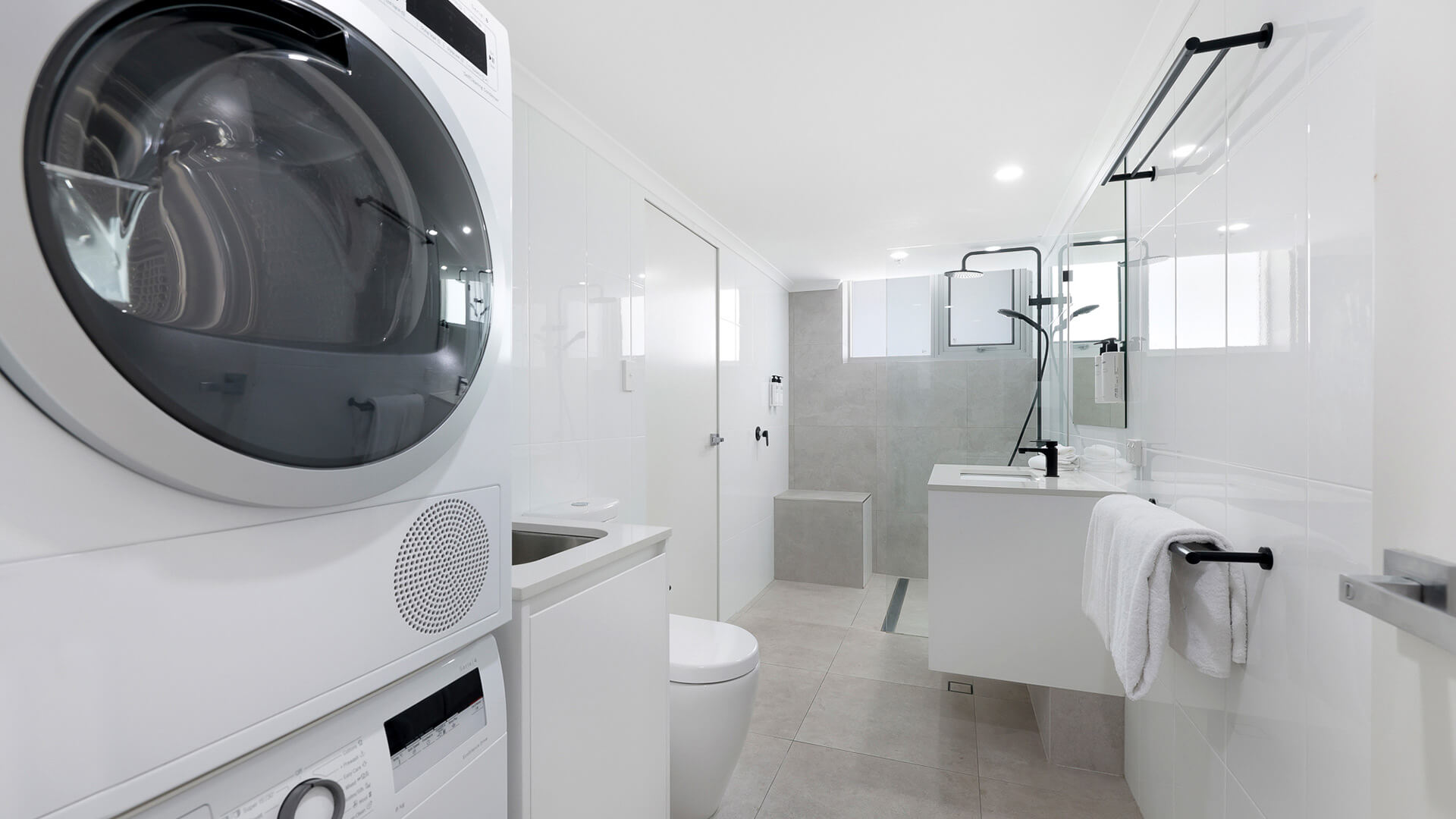 Modern bathroom and laundry combination, featuring a washer, dryer, walk-in shower, and vanity.
