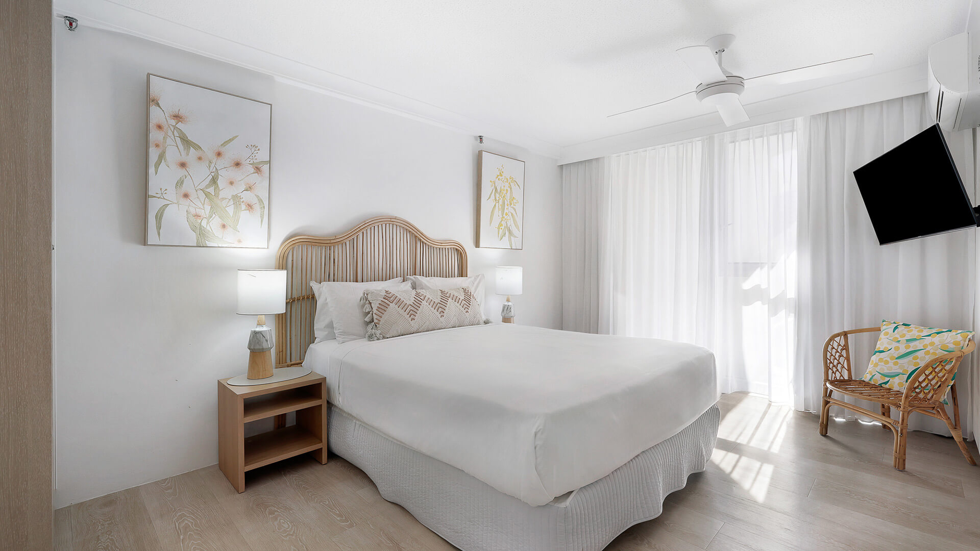 Coastal-style featuring a queen bed with rattan headboard, neutral tones, natural light, and a wall-mounted TV.