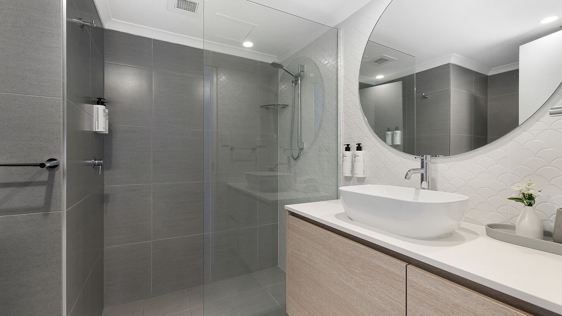 Modern bathroom featuring a walk-in rainfall shower, round mirror, vessel sink, and sleek white and grey finishes.