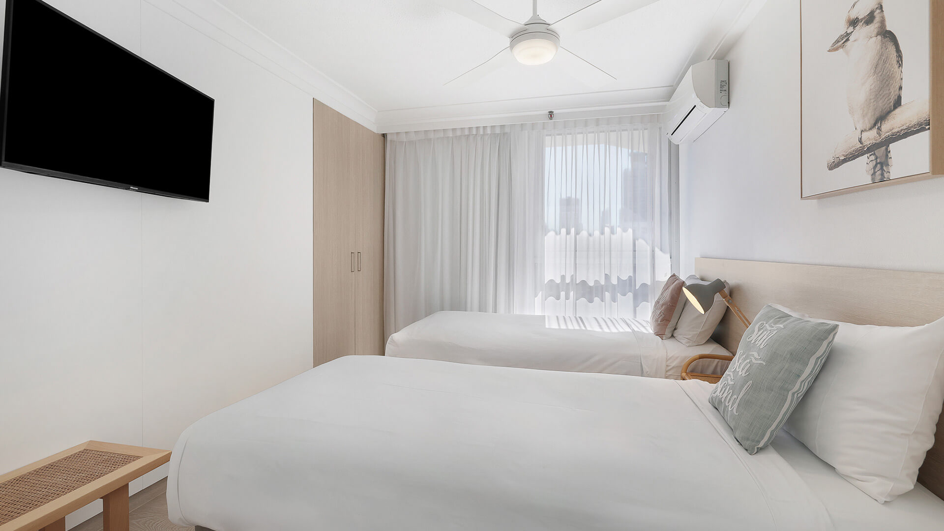 Second bedroom featuring one queen and one single bed, built-in wardrobe, ceiling fan, and wall-mounted TV in a bright and airy coastal setting.