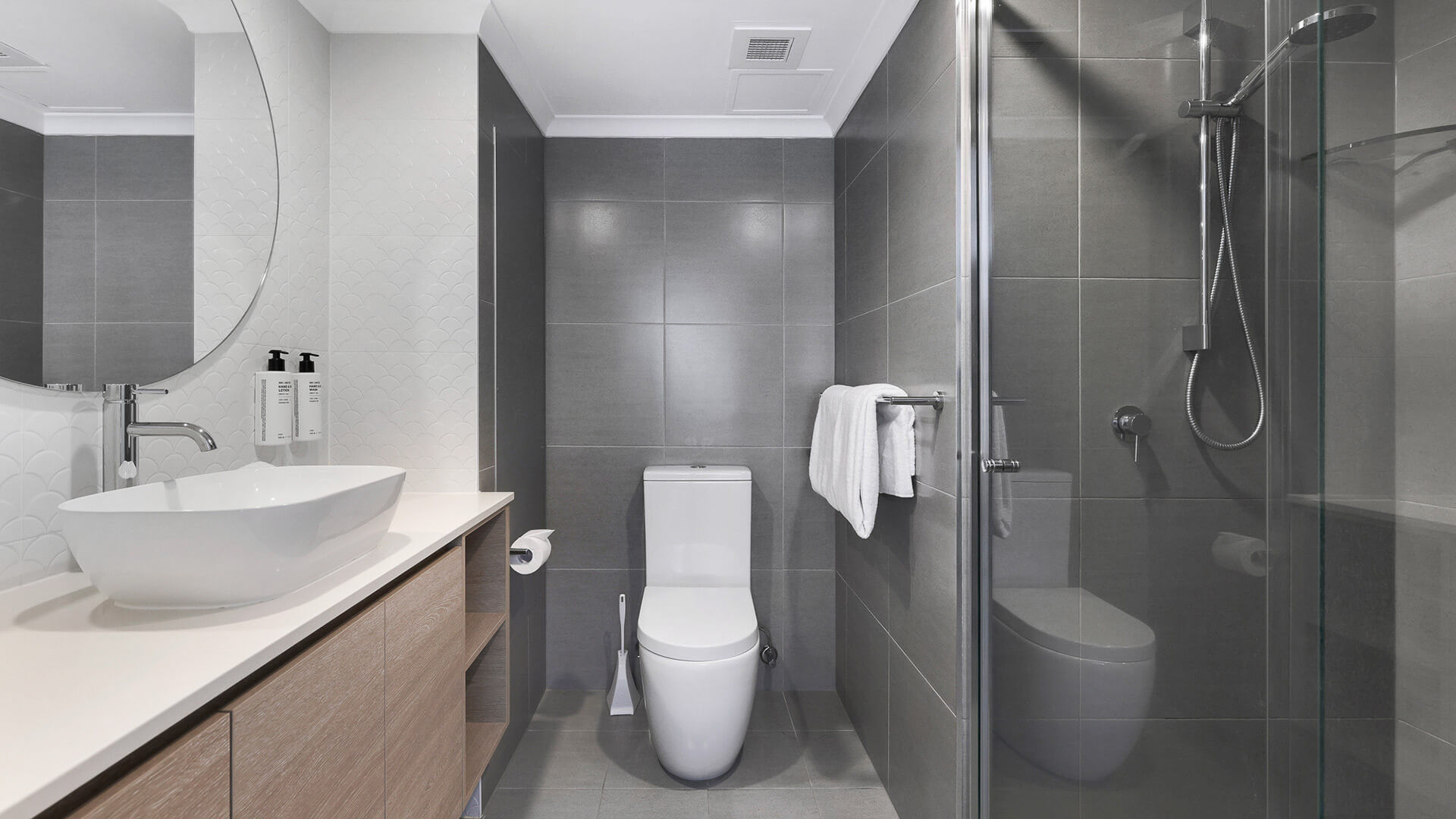 Modern bathroom featuring a walk-in glass shower, toilet, floating vanity with vessel sink, round mirror, and tiled walls in soft grey and white tones.