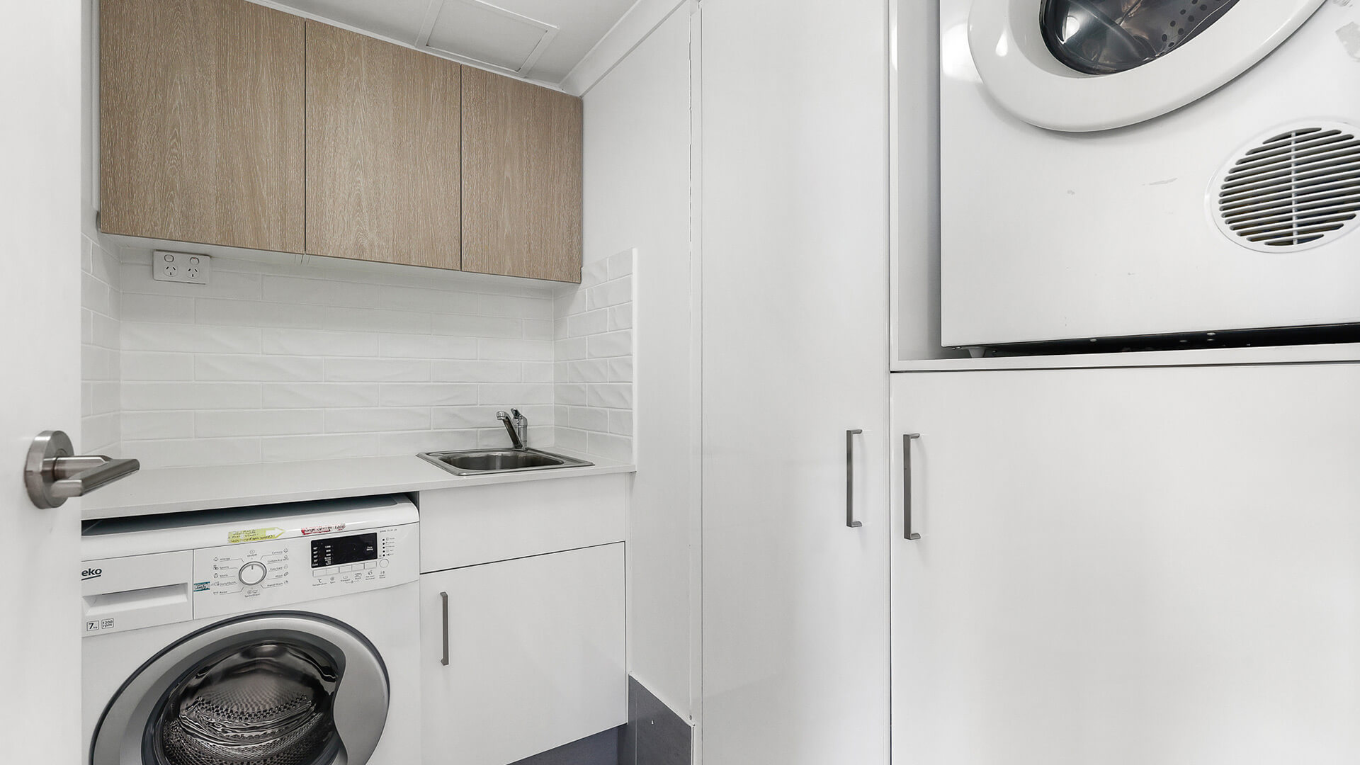 Fully equipped laundry featuring a front-loading washing machine, wall-mounted dryer, stainless steel sink, white cabinetry, and overhead timber-look cupboards.