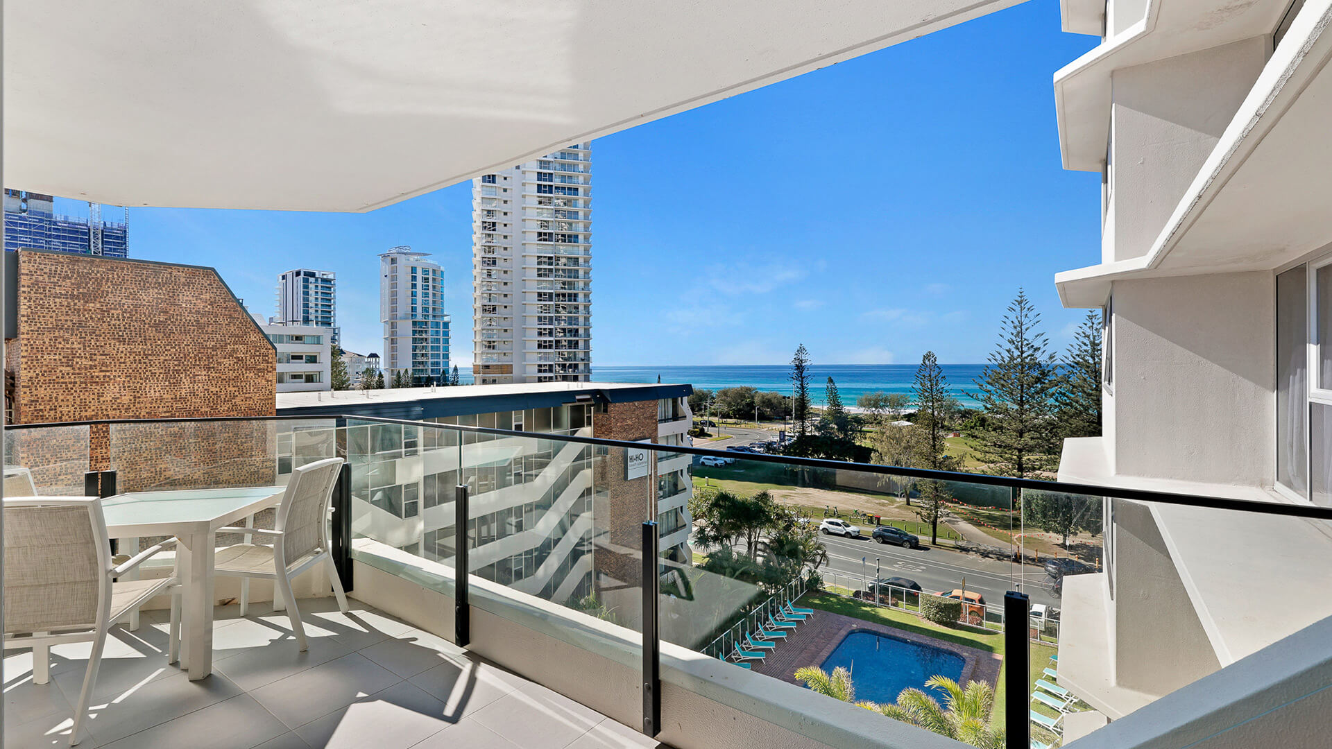 Private balcony overlooking the pool area with partial ocean views, an outdoor dining setting, and sights of Broadbeach high-rises and the coastline.