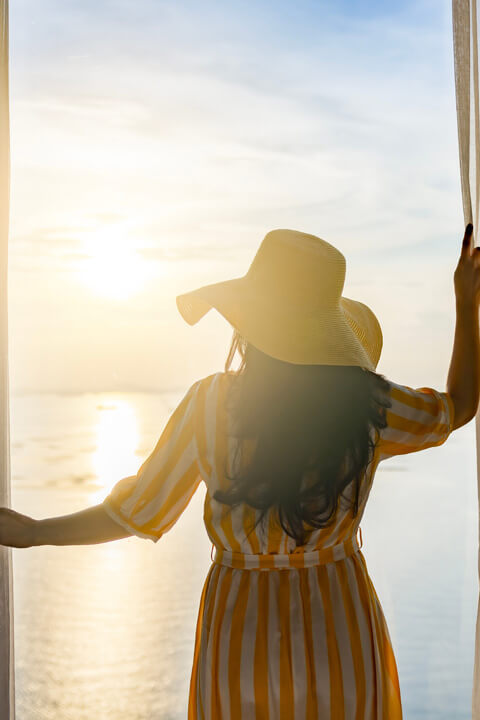 Woman in a sunhat opening curtains to a beautiful ocean sunrise, enjoying a relaxing coastal getaway.