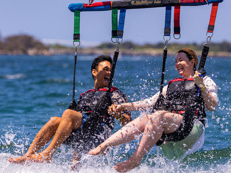 Couple parasailing over the ocean, laughing and holding hands as they skim the water's surface.