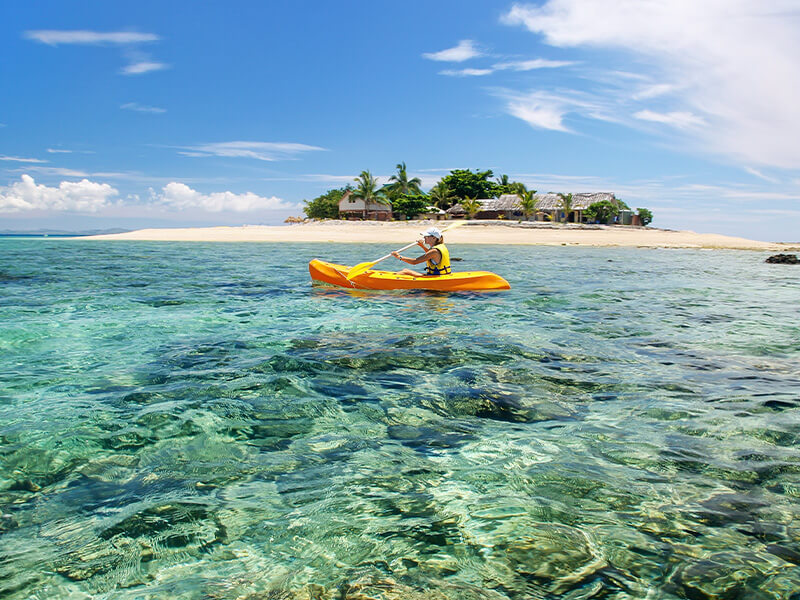 Person kayaking in clear turquoise waters near a tropical Fiji island.