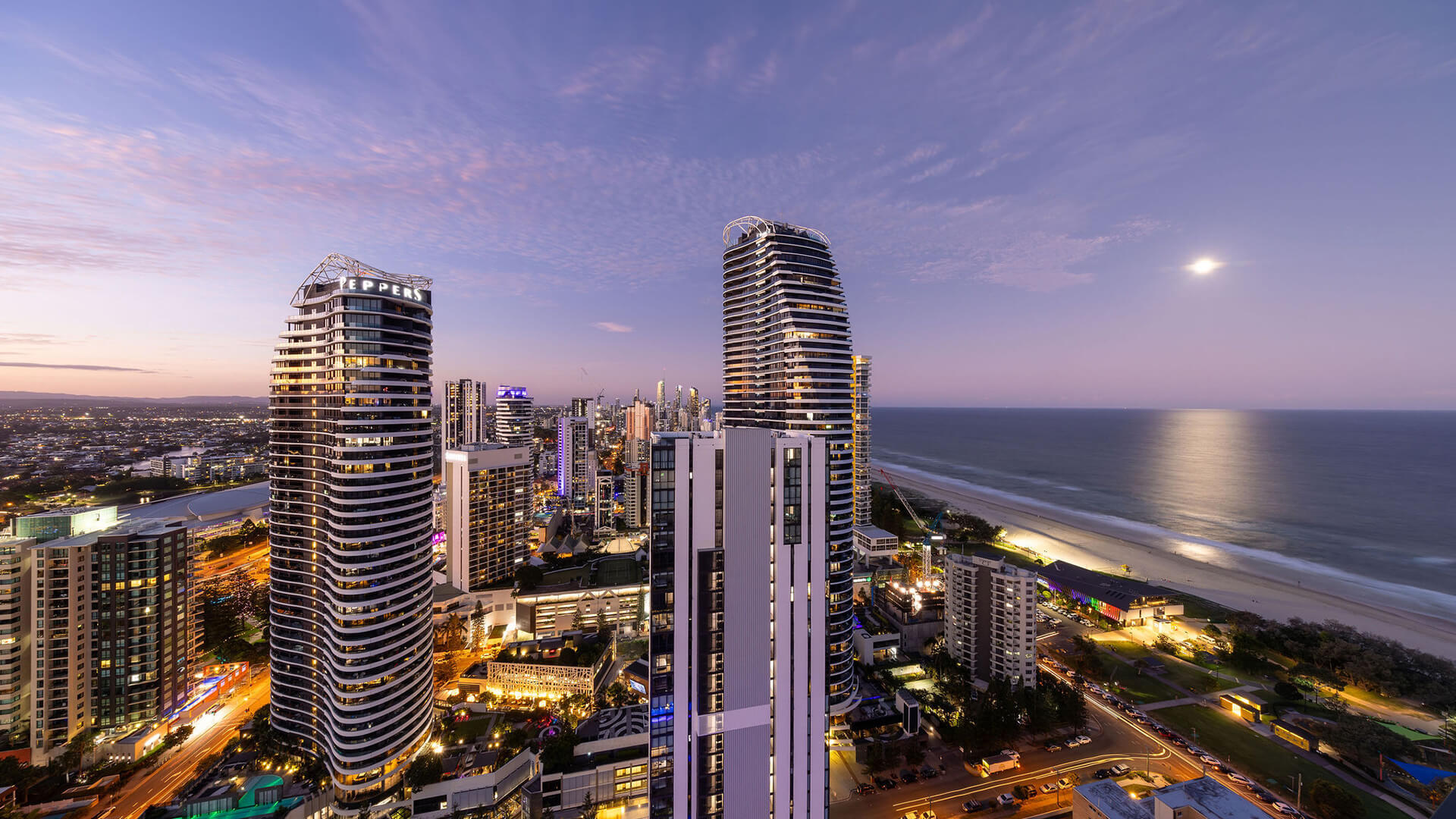 City skyline at dusk with illuminated high-rise buildings, a glowing moon over the ocean and reflections shimmering on the water.