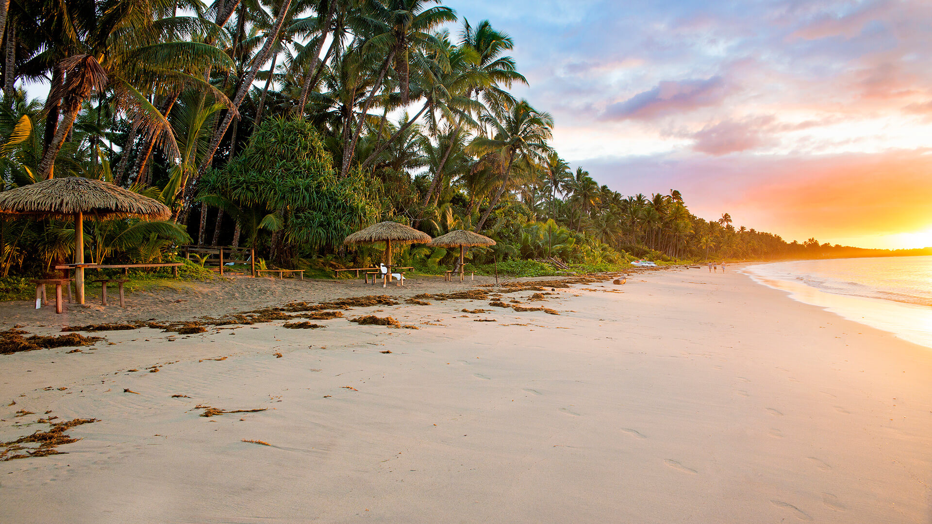 Golden sunset over a palm-fringed beach at ULTIQA Fiji Palms Beach Resort, with thatched umbrellas, picnic tables and gentle waves lapping the shore.