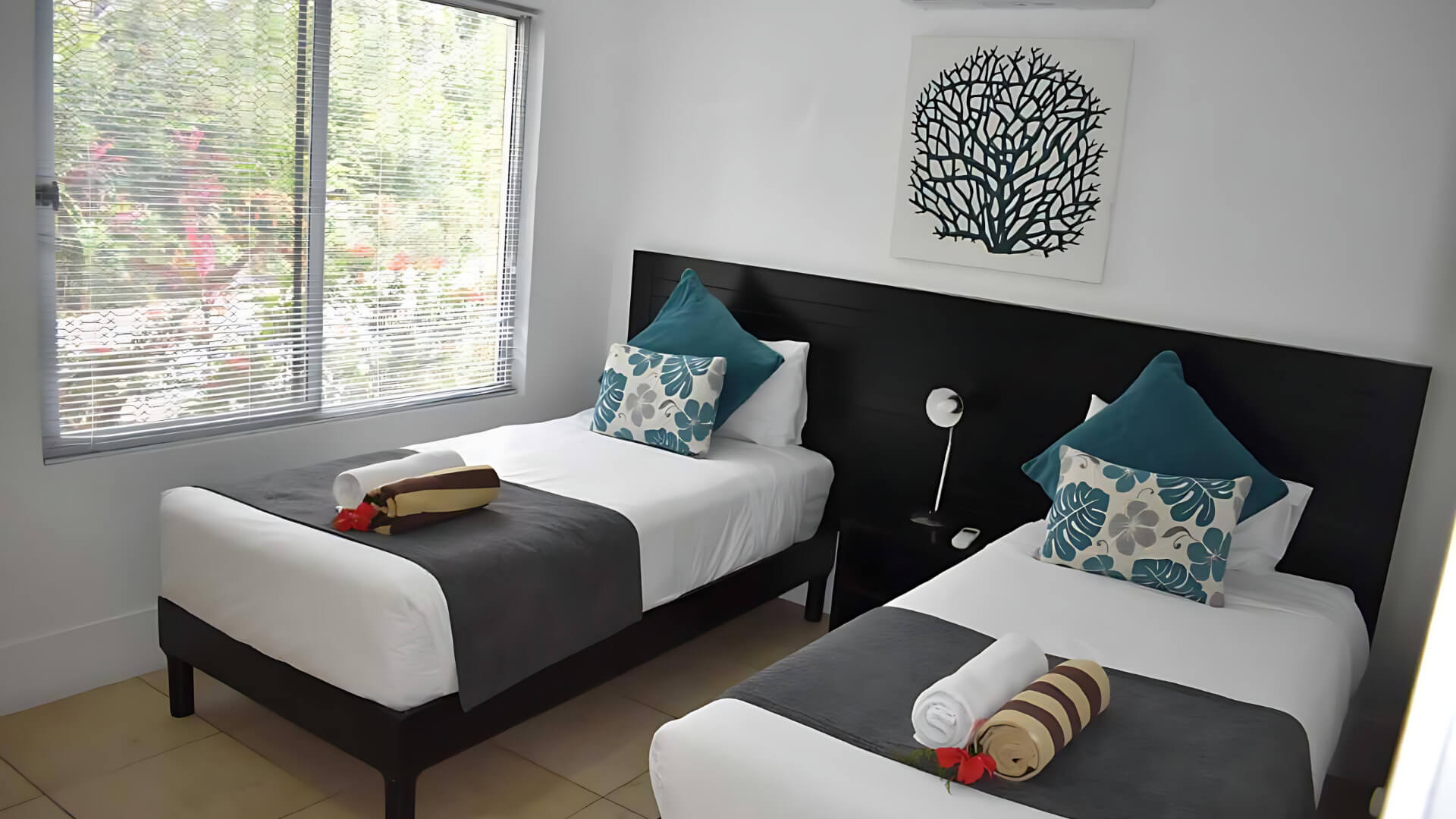 Twin bedroom in Standard 2 Bedroom 1 Bathroom Apartment at ULTIQA Fiji, featuring two single beds with grey throws, teal and patterned cushions, and a large window with garden views.