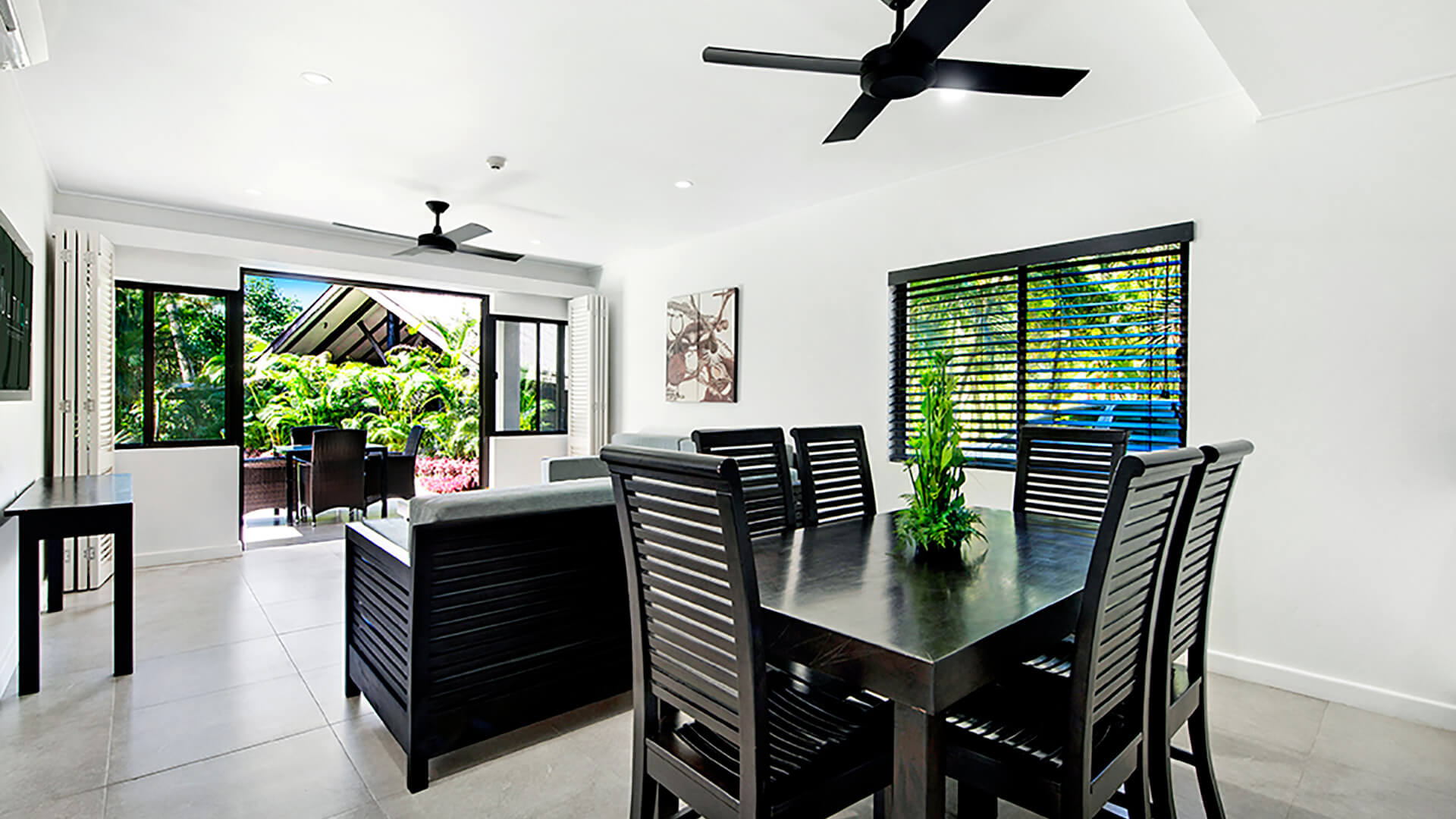 Spacious dining and living area featuring an 8-seater dining table, two sofas, and sliding doors opening to an outdoor dining space with tropical garden views.