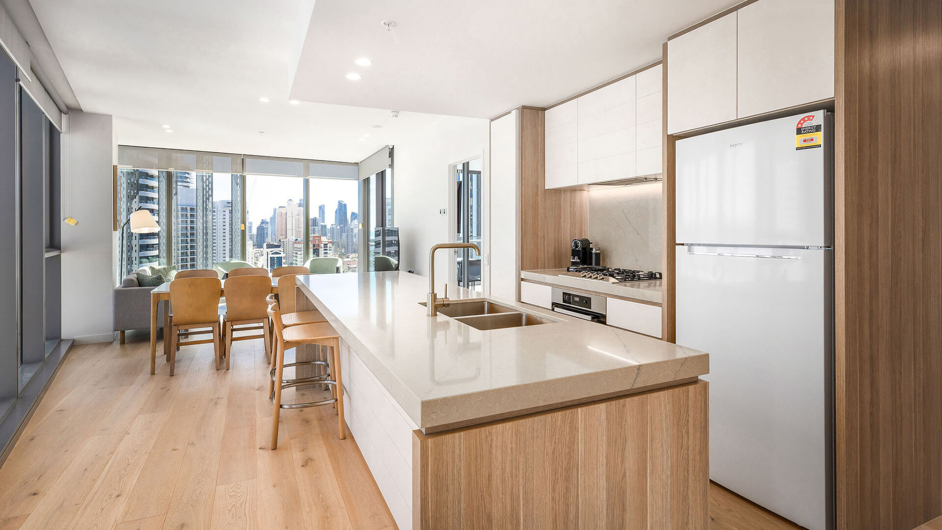 Modern kitchen in a 2 Bedroom, featuring a large island bench, built-in appliances, and wooden cabinetry with city views beyond.