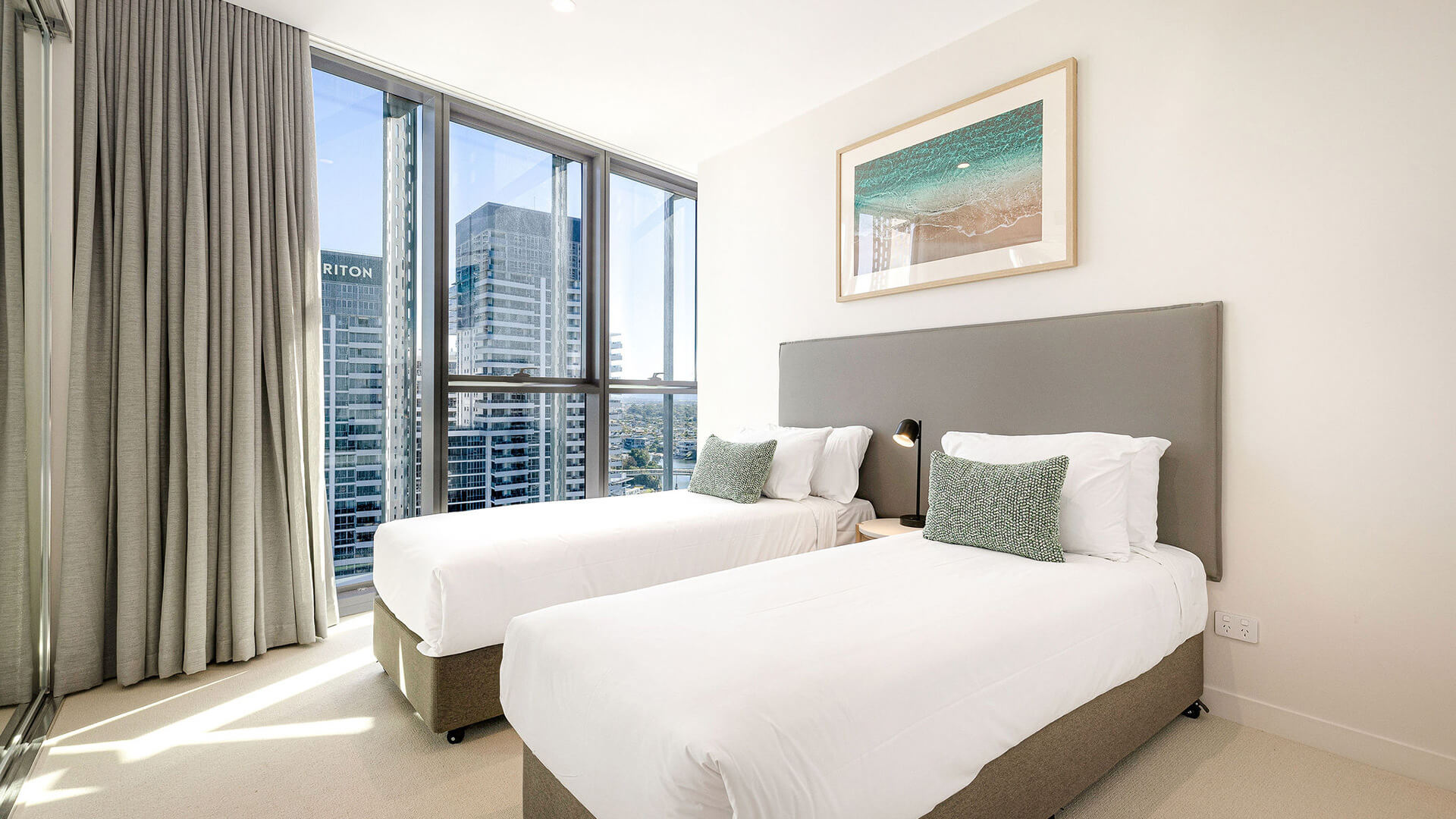 Twin bedroom in 3 Bedroom Suite at ULTIQA Signature, featuring two single beds, beach artwork, and city views through large windows.