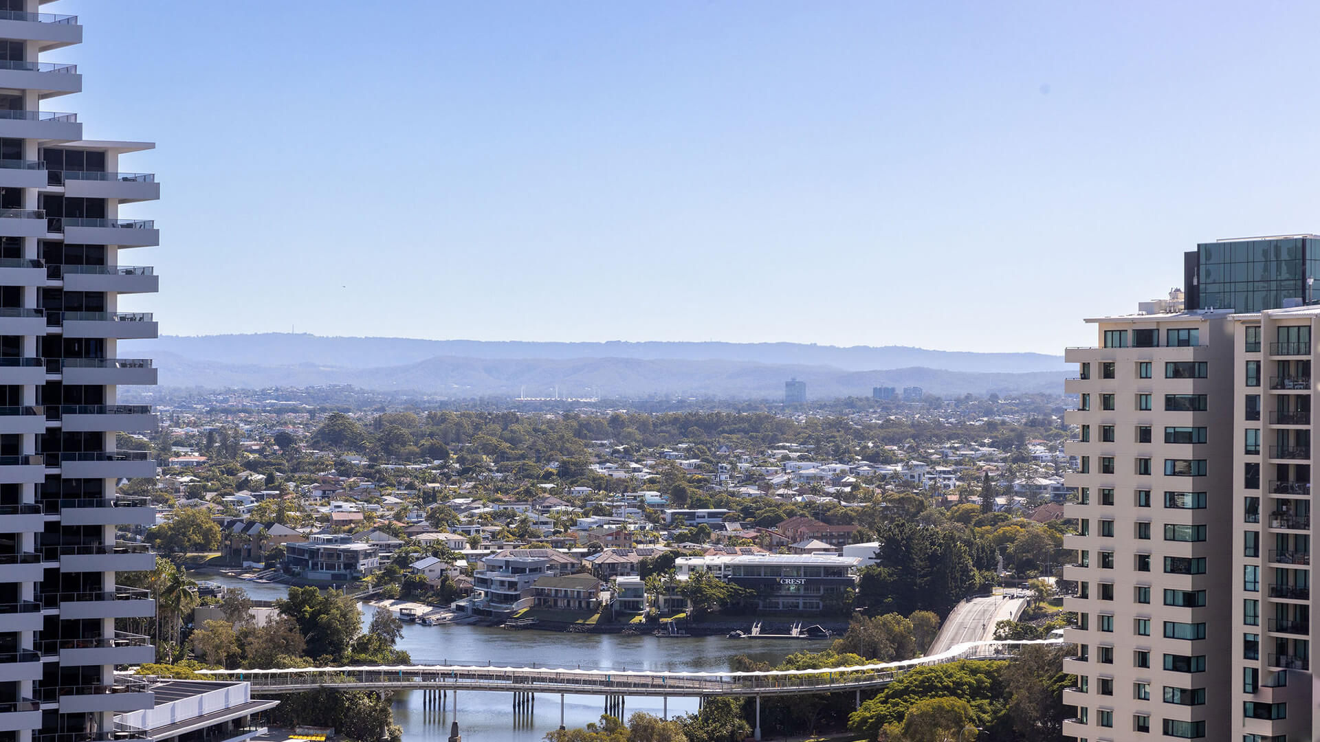 View from 3 Bedroom Suite at ULTIQA Signature, showing city skyline, river with pedestrian bridge, and distant hills under a clear blue sky.