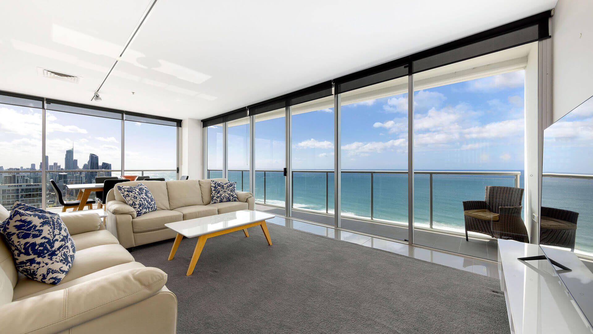 Living and dining area with beige sofas, white coffee table, and ocean views in ULTIQA Air’s 3 Bedroom Ocean View Suite. Balcony with wicker seating.